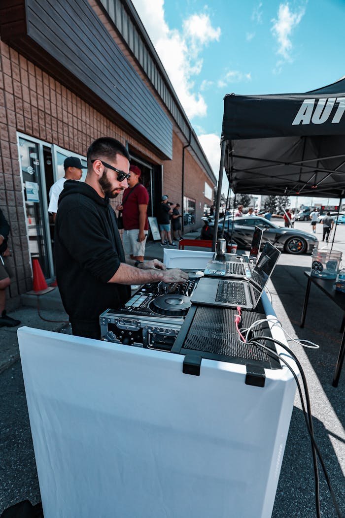DJ mixes music at an outdoor motoring event with laptops and sound mixer.